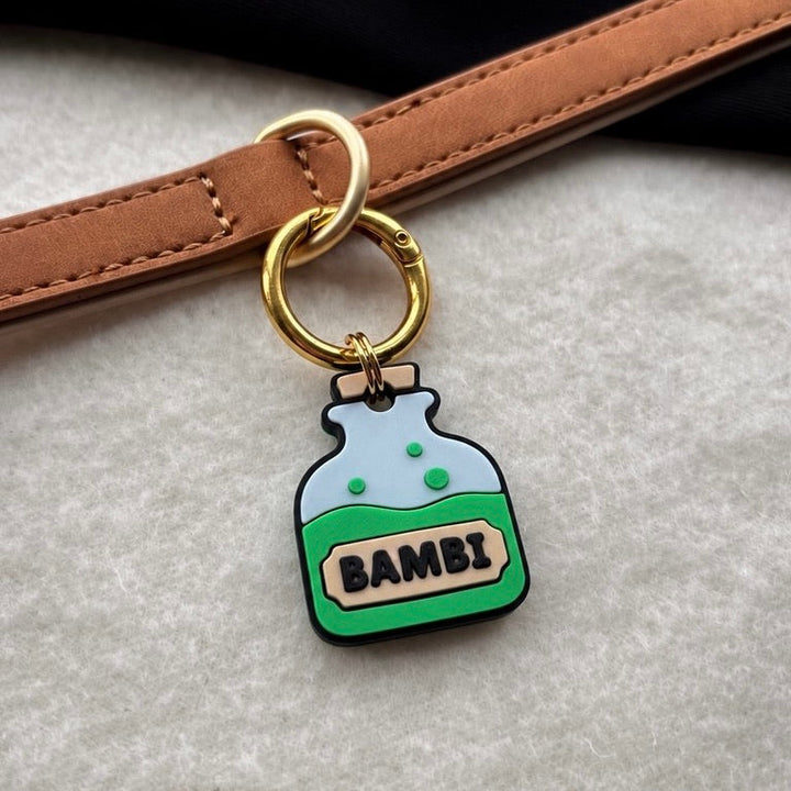 A close-up of a brown leather strap with a gold-tone key ring and a small charm shaped like a potion bottle labeled 'BAMBI'.