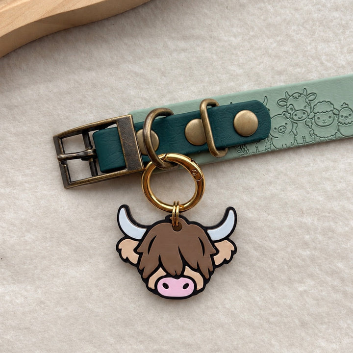A close-up of a pet collar with a turquoise leather strap, antique brass buckle, and a cute Highland cow tag hanging from a gold ring, displayed on a textured cream surface.