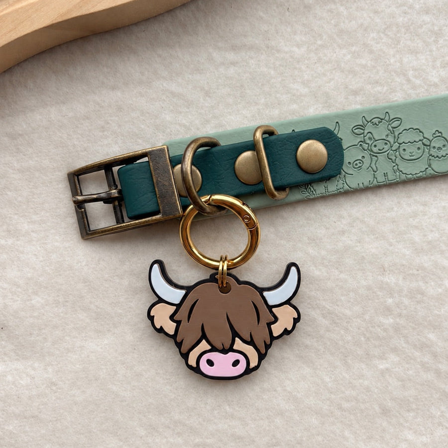 A close-up of a pet collar with a turquoise leather strap, antique brass buckle, and a cute Highland cow tag hanging from a gold ring, displayed on a textured cream surface.