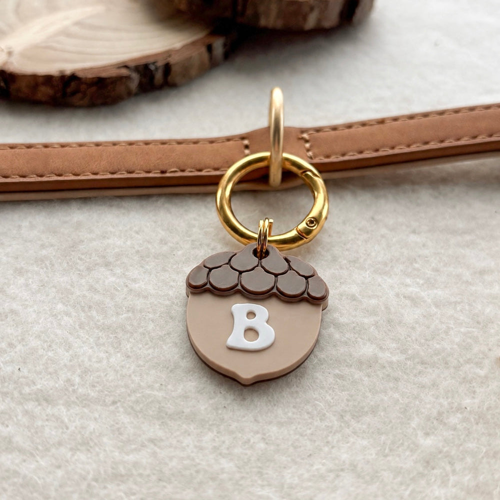A close-up of a tan leather strap with a gold ring and an acorn-shaped charm. The charm is tan with a white letter 'B' in the center and a dark brown textured top.