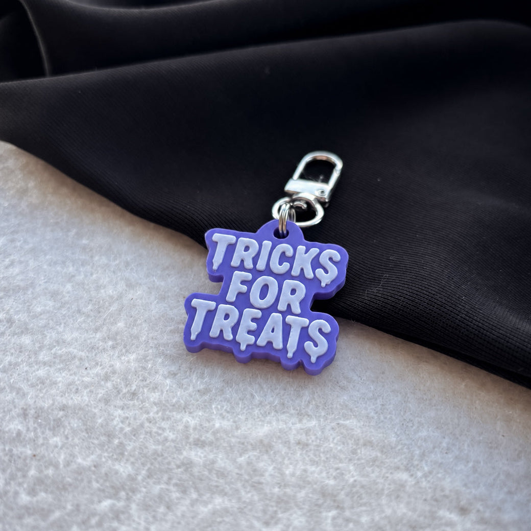 A small purple and white charm with the words 'TRICKS FOR TREATS' in raised white letters, attached to a silver clasp, resting on a black fabric surface over a light gray textured background.