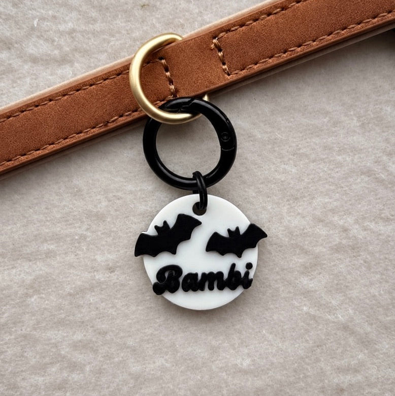 A close-up of a brown leather strap with a gold-tone ring, attached to a black ring holding a round white charm with black bat silhouettes and the name 'Bambi' in script.