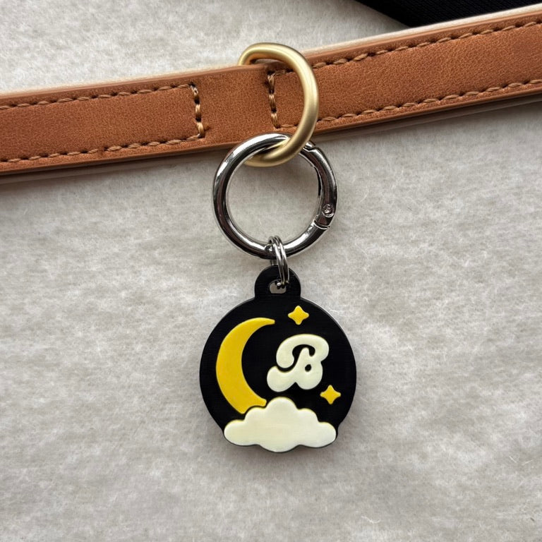A close-up of a circular black charm keyring with a yellow crescent moon, white cloud, and a stylized white letter B, attached to a tan leather strap with a gold-tone ring.