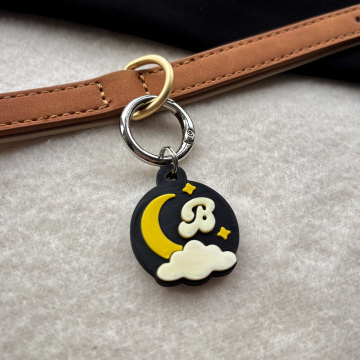 A close-up of a circular black charm keyring with a yellow crescent moon, cream cloud, and a stylized cream letter B on a tan leather strap against a light gray textured background.