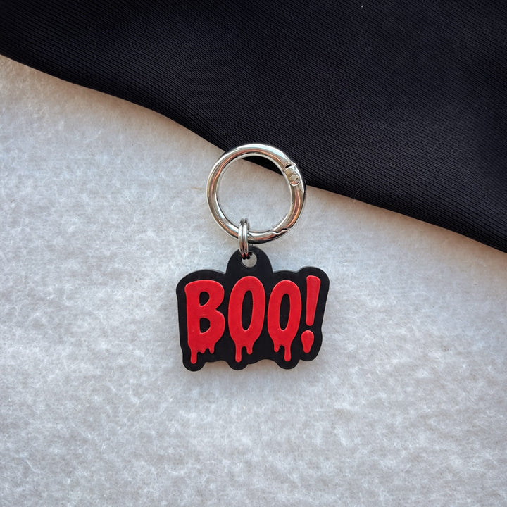 A Halloween-themed dog tag featuring a red dripping 'BOO!' text charm with a black outline and a silver metal key ring, displayed on a split white textured surface and black fabric background.