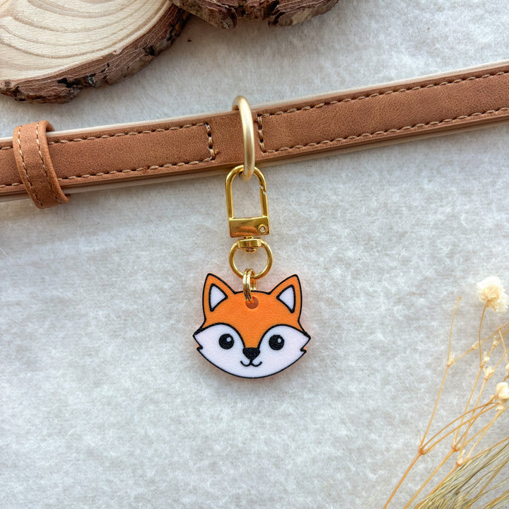 A small fox head charm with a gold clasp hanging from a tan leather strap, styled on a light textured surface with wood slices and dried grass.