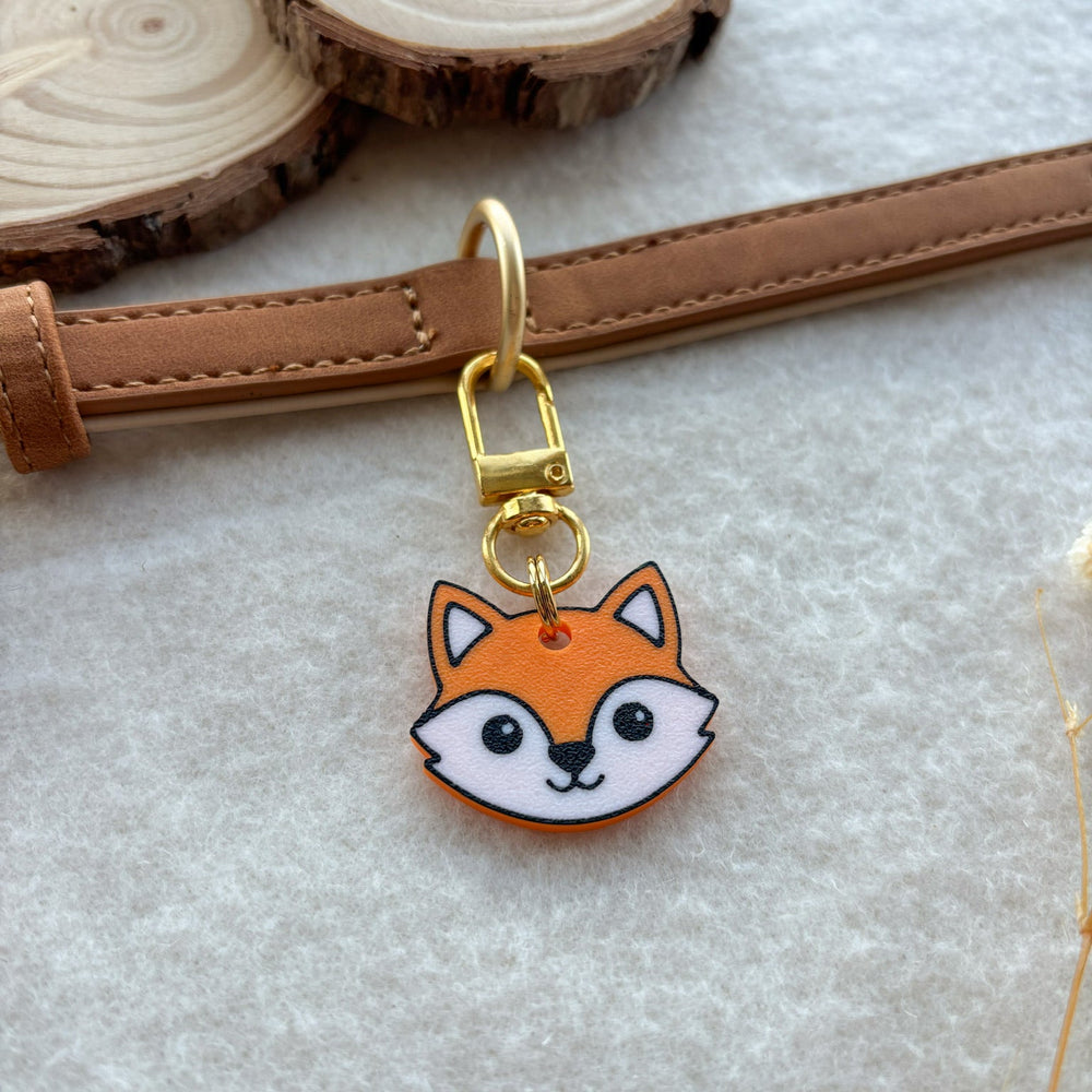 A small fox-face charm with a gold clasp and ring, hanging from a tan leather strap on a light fabric surface, with wood slice props in the background.