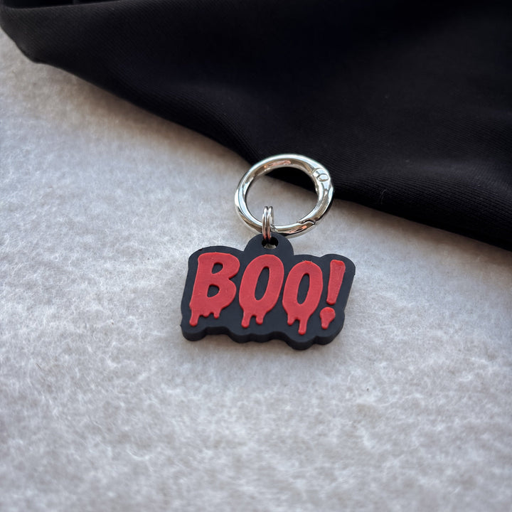 A Halloween-themed keychain charm featuring the word 'BOO!' in dripping red letters on a black background, attached to a silver metal key ring, resting on a textured light gray surface with a black fabric backdrop.