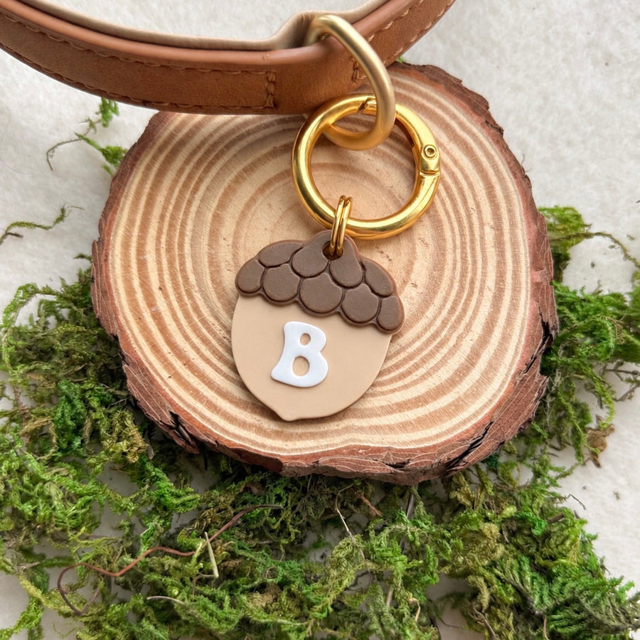 A close-up of a personalized dog collar with a tan leather strap and a gold clasp, featuring an acorn-shaped charm with a light tan center, dark brown textured top, and a white letter 'B'. The charm is displayed on a rustic wood slice surrounded by green moss.