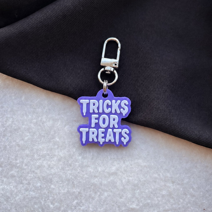 A small purple  'TRICKS FOR TREATS' dog tag with a silver lobster clasp, resting on a black fabric surface over a light gray textured background.