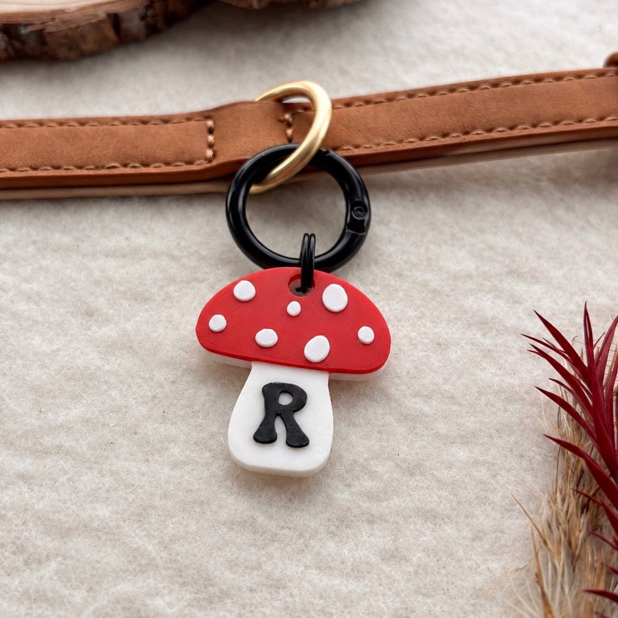 A mushroom-shaped charm with a red cap, white spots, and a white stem featuring a black letter 'R', attached to a black ring and gold-tone hardware, shown on a tan leather strap against a neutral textured background with wood slices and dried grass.
