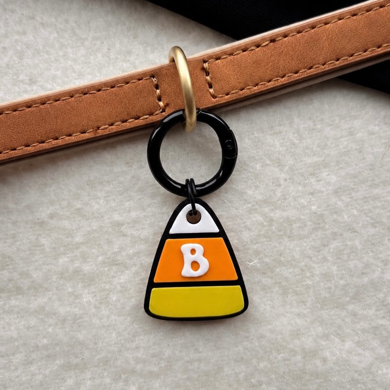 A close-up of a tan leather belt with a black ring and a candy corn-shaped charm featuring orange, yellow, and white sections with a white letter 'B' in the center.