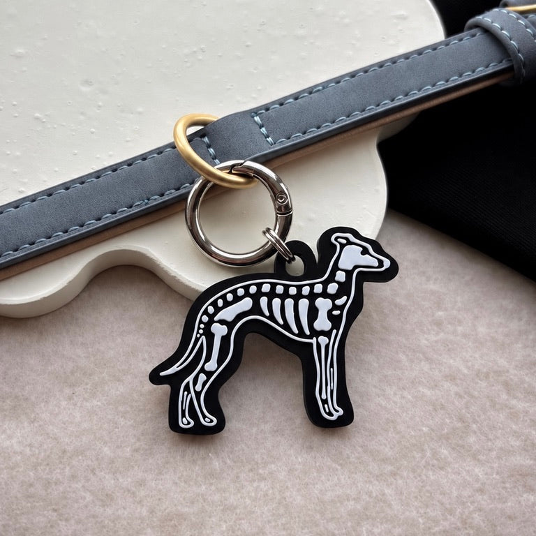 A black and white dog skeleton charm keychain with gold-tone hardware, shown hanging from a gray leather strap on a textured beige surface.