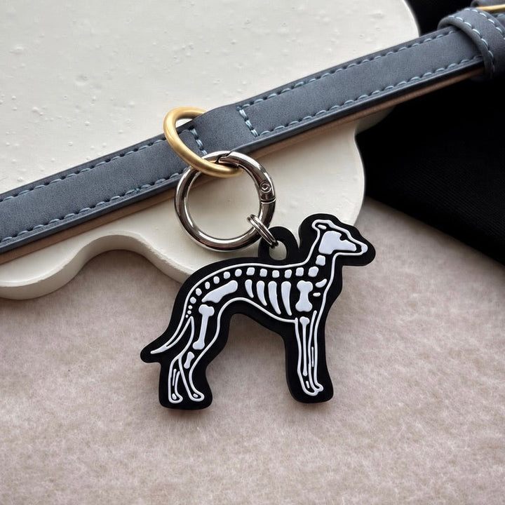 A black and white dog skeleton charm keychain with gold-tone hardware, shown hanging from a gray leather strap on a textured beige surface.