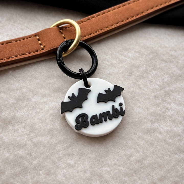 A close-up of a circular white charm with black bat silhouettes and the name 'Bambi' in black script, attached to a black ring and gold-tone hardware, resting against a tan leather strap on a light textured surface.