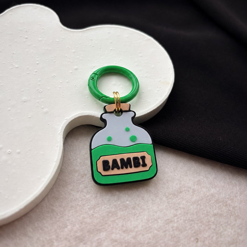 A small charm shaped like a green potion bottle with the word 'BAMBI' on the front, attached to a green ring. The charm is styled on a white sculptural prop with a black fabric background.