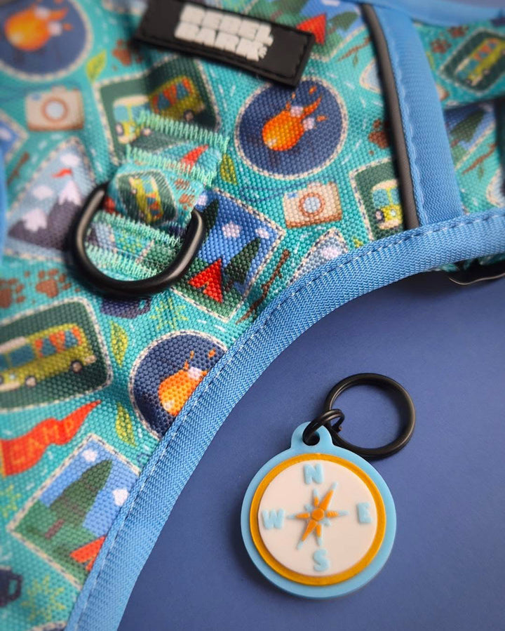 A close-up of a blue dog harness with a colorful cartoon print, featuring a black metal D-ring and a round blue and white compass-shaped tag with a black key ring.