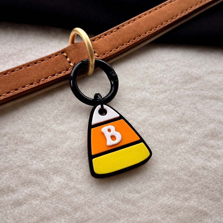 A close-up of a black ring charm shaped like a candy corn, featuring horizontal stripes of white, orange, and yellow with a white letter 'B' in the center, hanging from a brown leather strap on a textured beige surface.