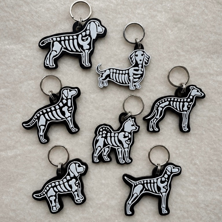 Seven dog skeleton dog tags in various breeds, featuring black and white dog skeleton designs on a textured beige background.