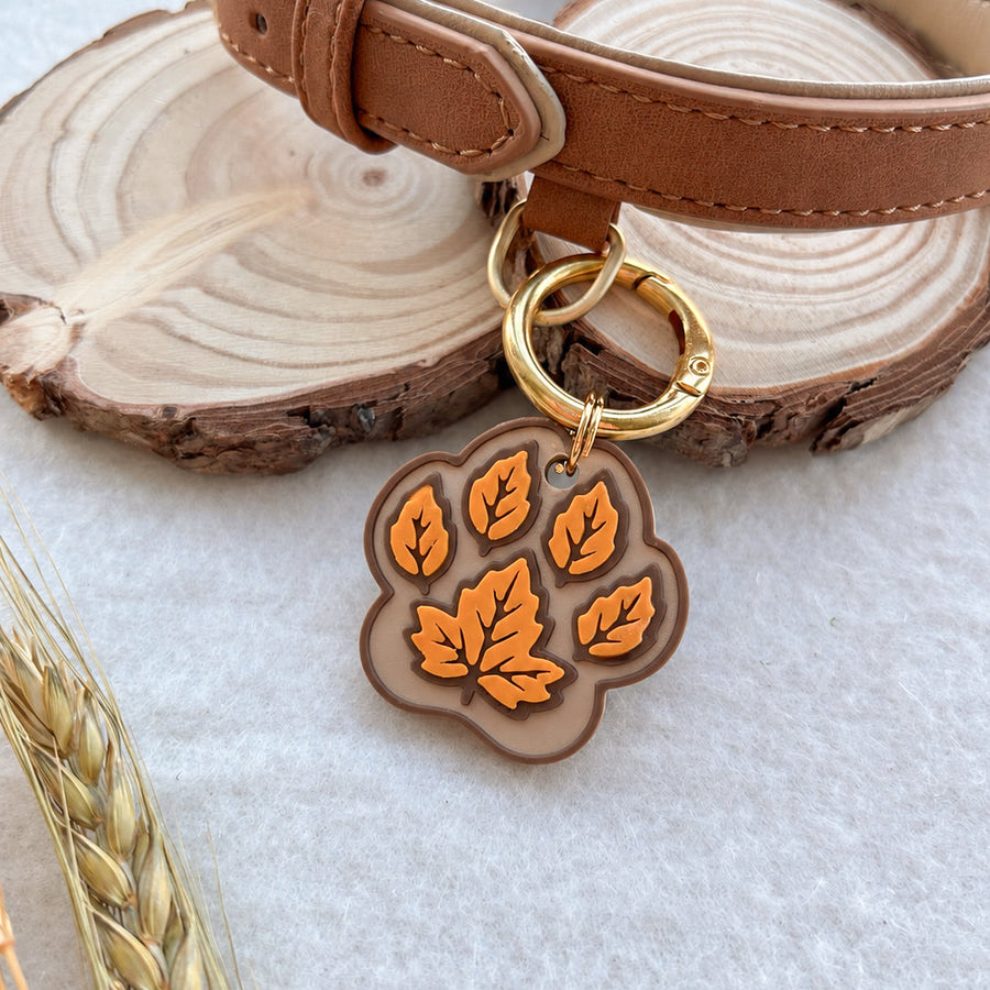 A tan leather pet collar with a gold-tone ring and a paw-print shaped tag featuring orange autumn leaves, displayed on rustic wood slices and wheat stalks.