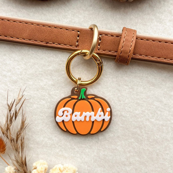 A close-up of a brown leather strap with a gold-tone ring and a pumpkin-shaped charm. The charm is orange with a green stem and the name 'Bambi' written across it in white script.